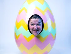 A person's face emerging from the opening of an oversized painted Easter egg costume decorated with zigzag stripes in yellow, pink, and mint green, photographed against a soft white studio backdrop. Even, cool-toned studio lighting illuminates the egg surface from directly above, highlighting the glossy painted texture. The face is centered in the oval cutout, expression amused. Style: clean product-style holiday portrait. Mood: whimsical and bright.